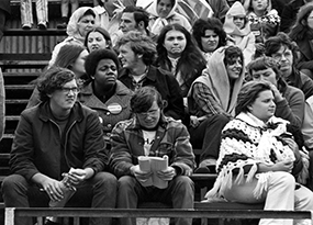 A crowd of young men and women wrapped in shawls and coats are sitting in the stands, watching a sport event.  One woman wears a “Manhattan” pin prominently on her coat. 